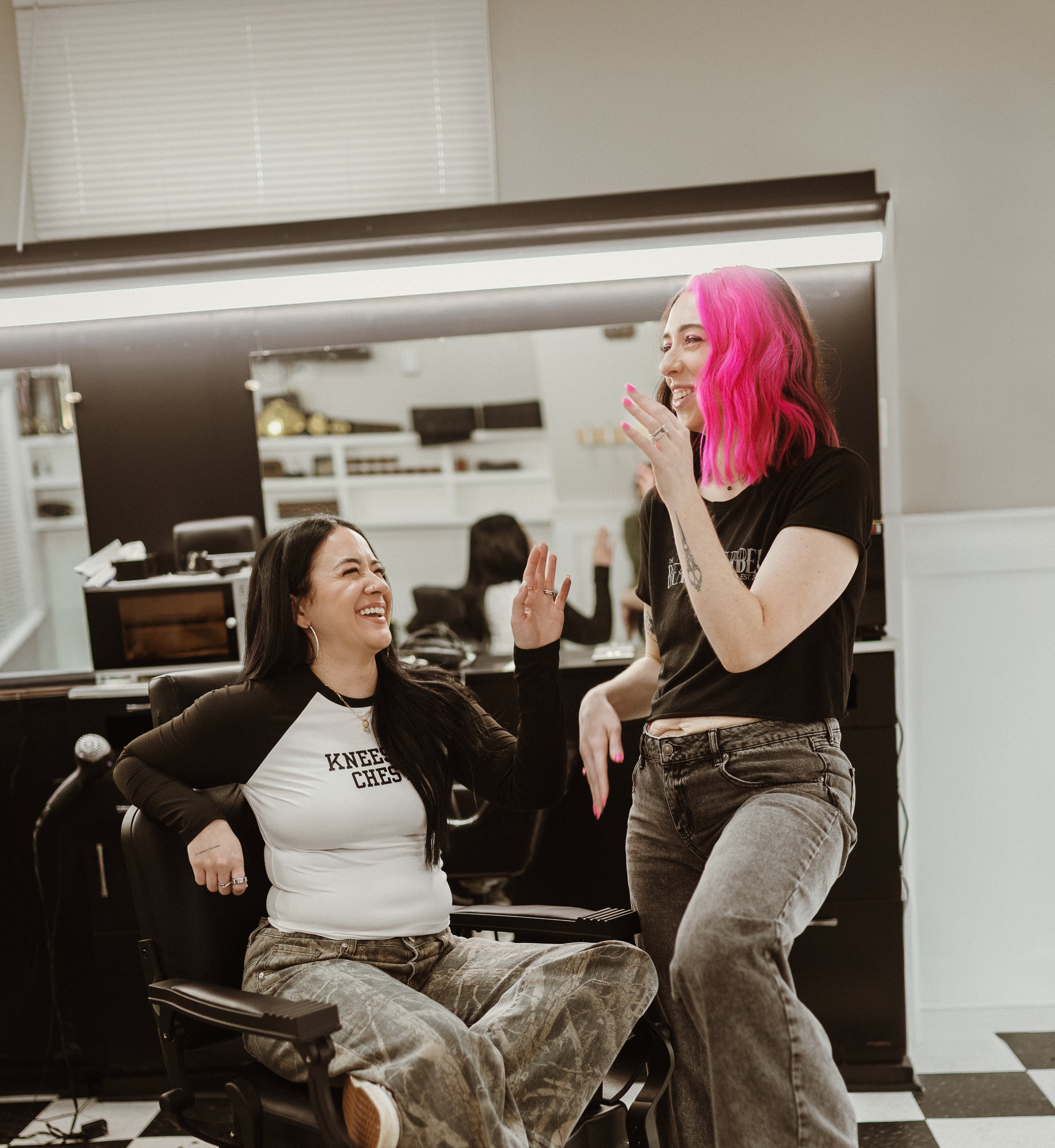 Two women in a salon setting, one with pink hair, in a checkered floor.